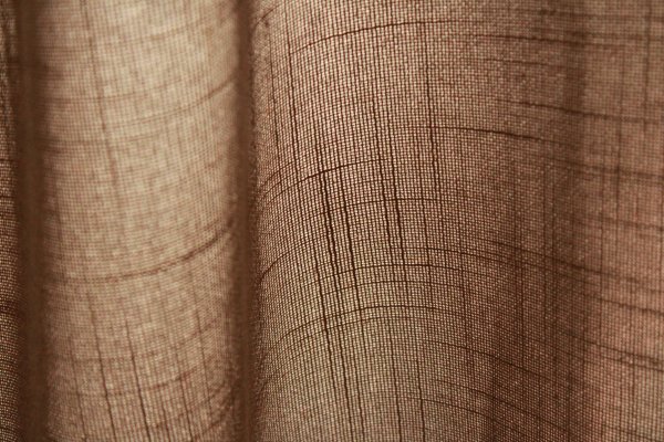 Stofstructuur van taupekleurige inbetween gordijnen met linnen look uit de Saphira Light collectie van Okika, lichtdoorlatend met zichtbare natuurlijke vezelstructuur.