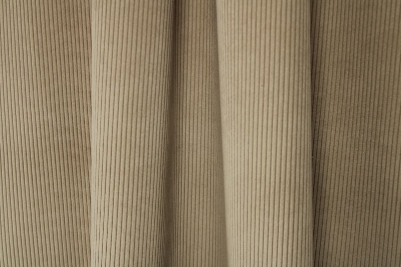 Sfeerfoto van softtaupe ribstof gordijnen uit de SYM2600 collectie van Okika, waarbij de verticale structuur van de ribstof zichtbaar is en de gordijnen in plooien zijn gefotografeerd